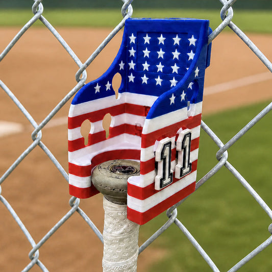 Bat Saver™ Patriotic Edition with jersey numbers in the charm slots attached to a chain-link fence whilst cradling a softball bat.